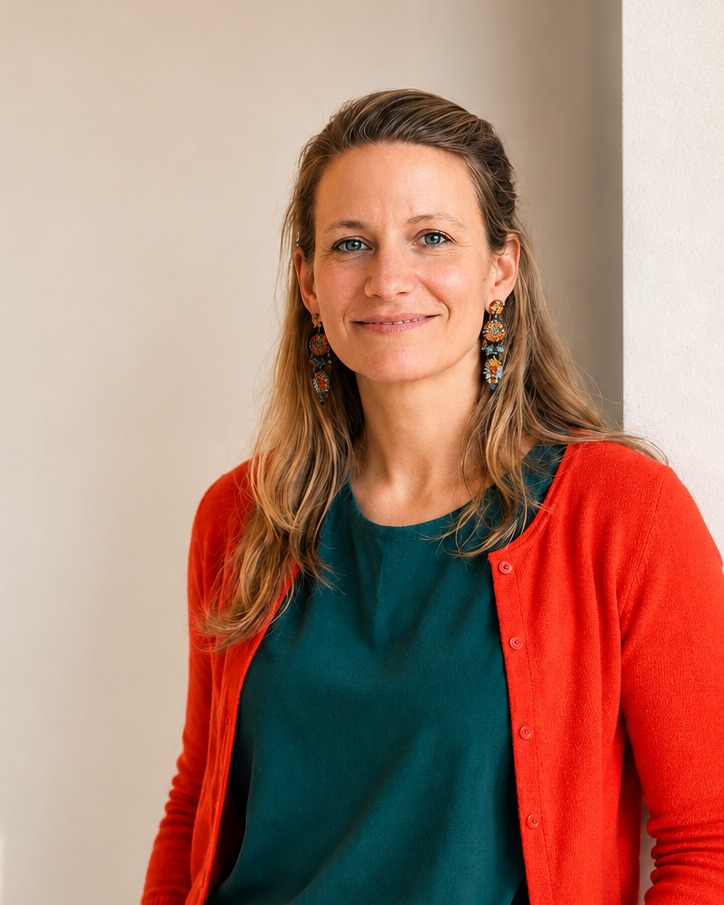 Stephanie Wille lehnt lächelnd an einer hellen Wand, in orangefarbener Strickjacke